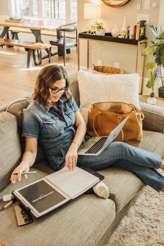 Adult woman in stylish denim outfit working at home on a couch with a laptop and planner, enjoying a productive day.